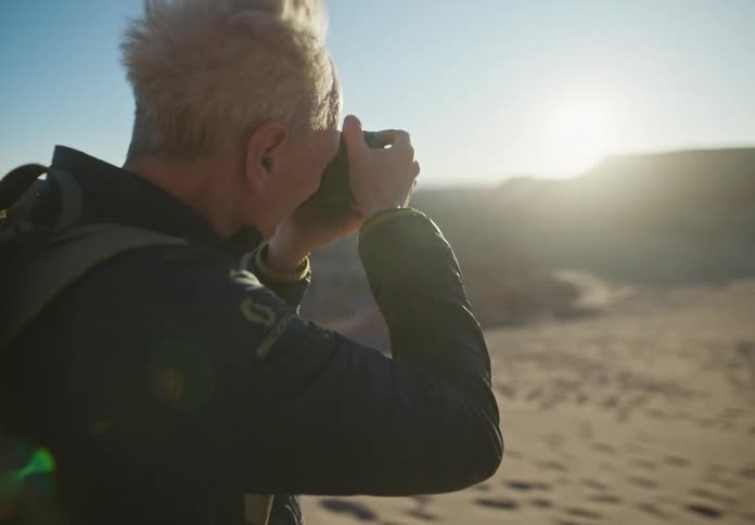 Man holding a camera and taking photos in an environment with desert dunes and mountains