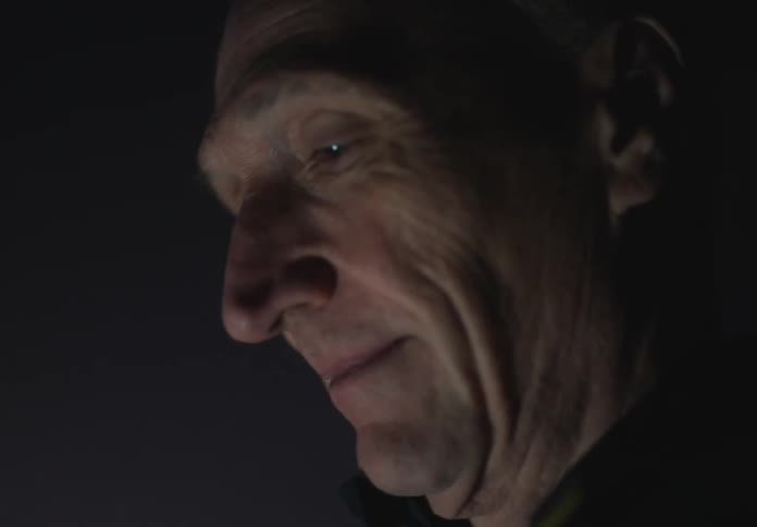 An older white man smirks and looks around him