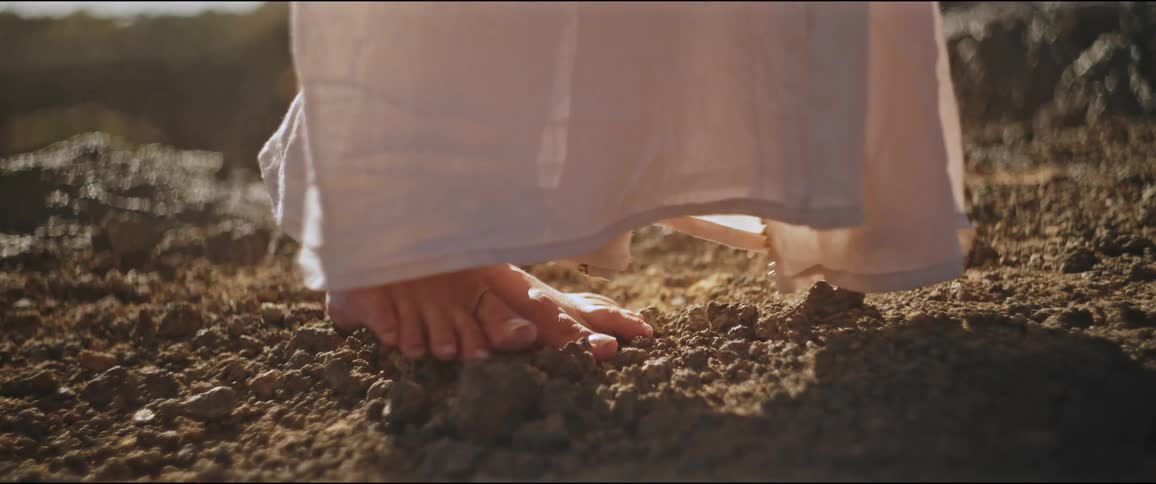 Closeup of a womans bare feet walking on rocky ground in a long white dress