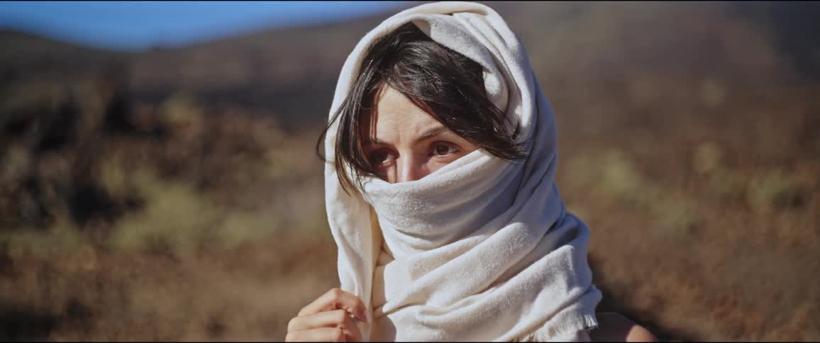 Young woman with a scarf covering her face with only her eyes visible while standing in a natural environment surrounded by mountains in the blurred background