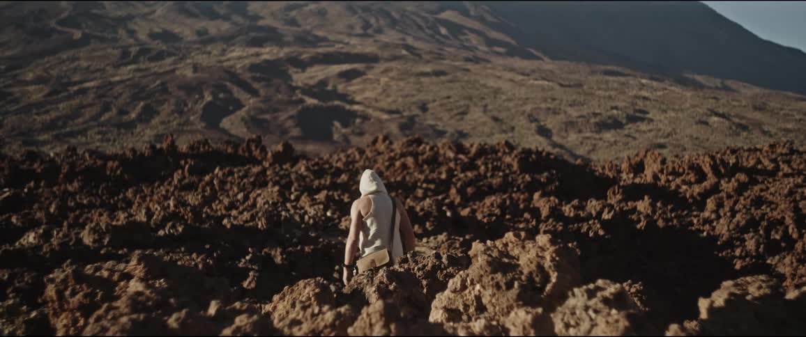 Person from behind wearing a light suit and walking on rocky and uneven terrain with mountains in the background under a clear sky