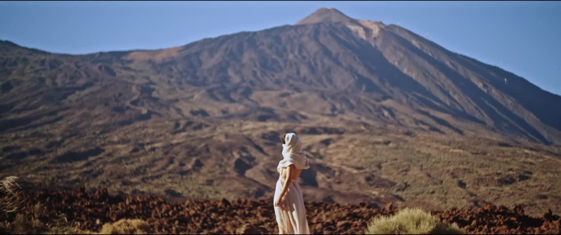 A woman in loose white clothes protecting her skin from the sun in a desert region with a large mountain in the background