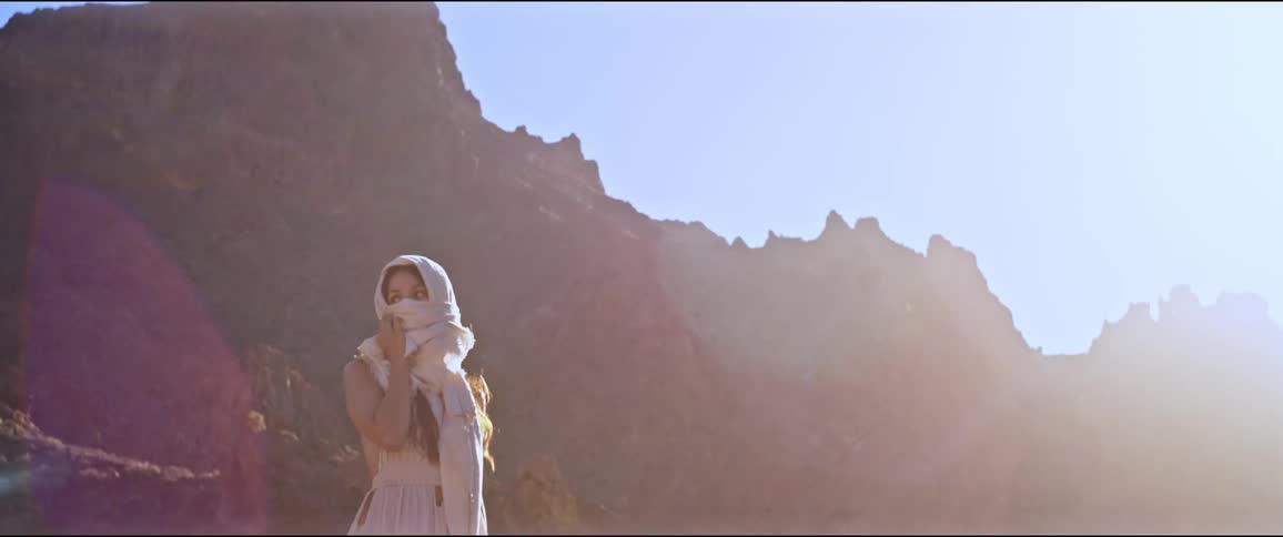 A woman wearing a scarf over her head stands in front of a mountain under a blue sky in a sunny day