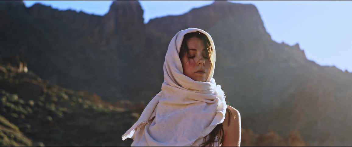 A woman wearing a scarf over her head stands in front of a mountain under a blue sky in a sunny day