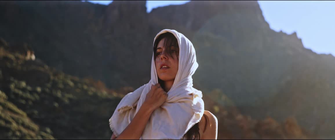 A woman wearing a scarf over her head stands in front of a mountain under a blue sky in a sunny day