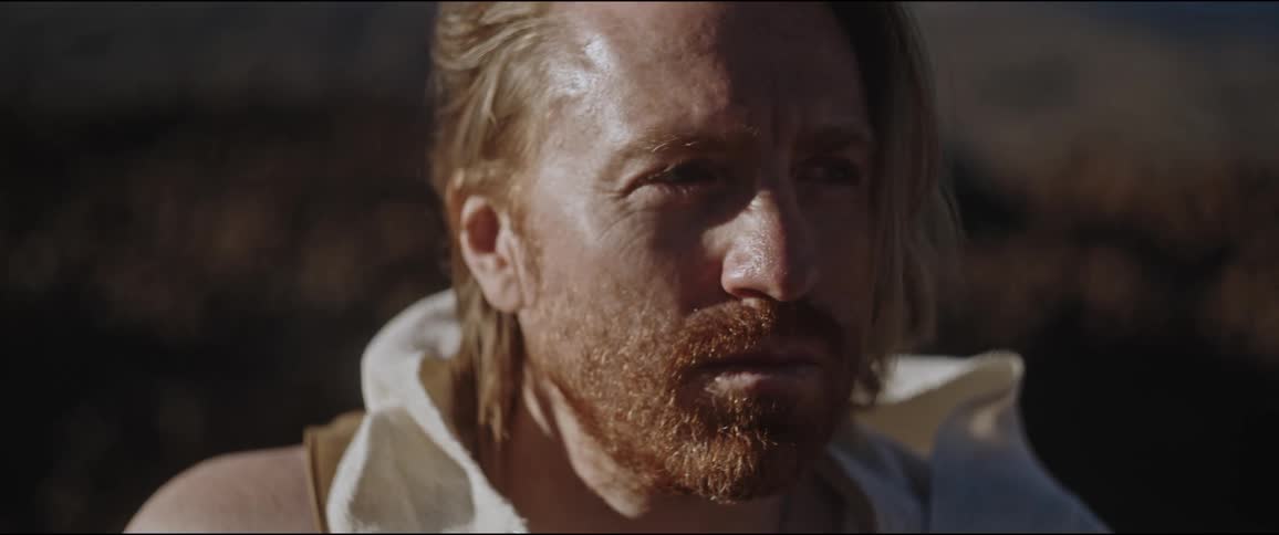 A bearded man with long hair wearing a sleeveless hoodie in nature