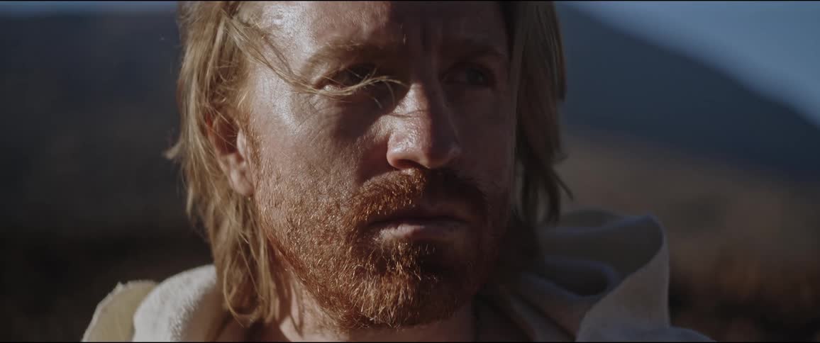 A bearded man with long hair wearing a sleeveless hoodie in front of a mountain