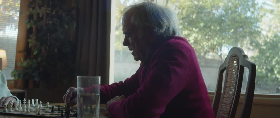 An elderly man sitting on a table at home playing chess near a window