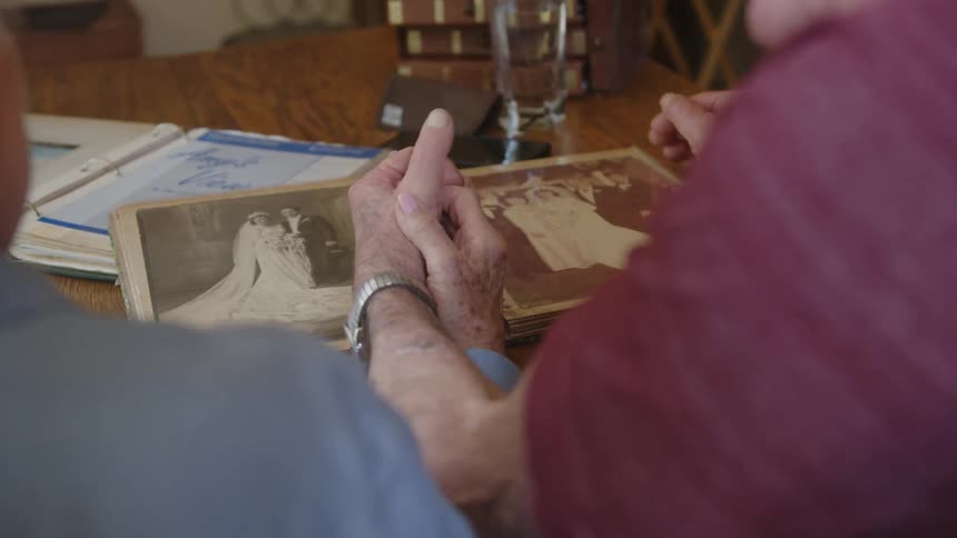 Elderly couple sitting at a table with their hands together looking at wedding photos in an album