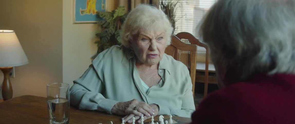 An elderly woman talking to an elderly man who is in front of her and in front of them there is a table with a game of chess