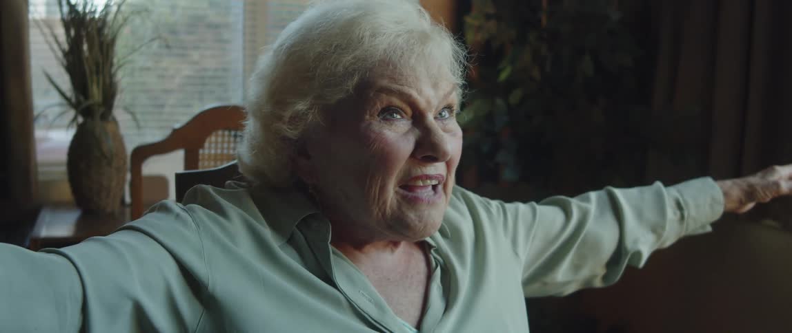 An elderly woman sitting in a chair in a room excitedly speaks