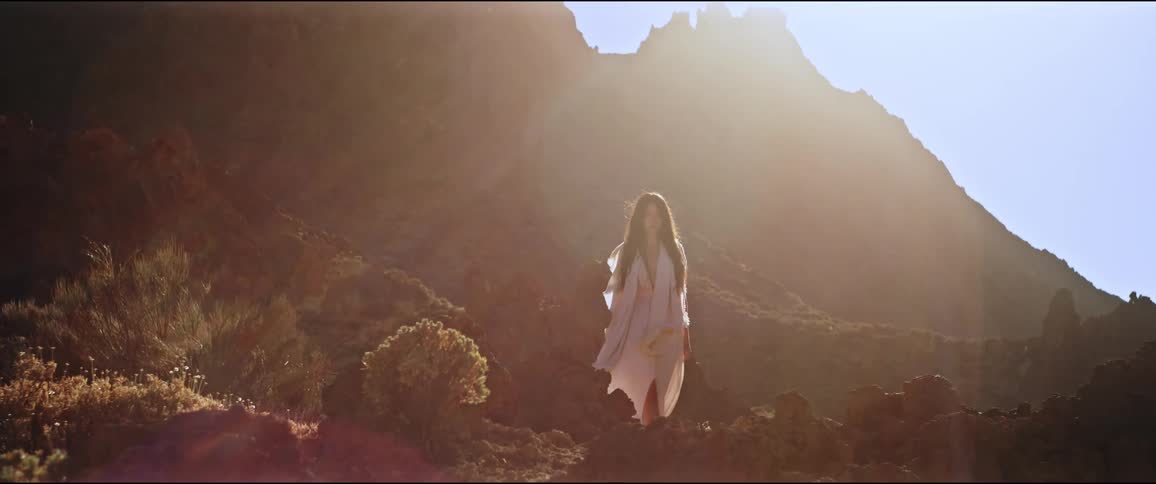 Woman in a white dress walking on rocky terrain backlit by the sun