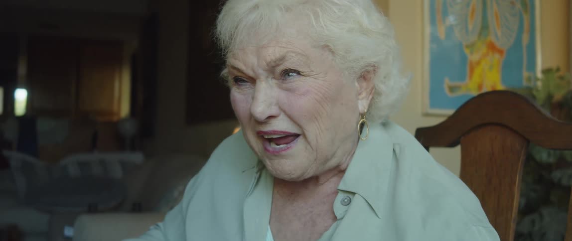 An elderly woman laughs and speaks while sitting in a dining room chair