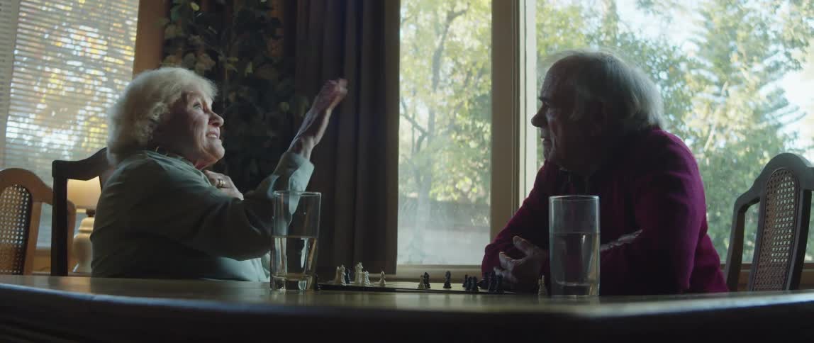 Two elderly people sitting at a table with the lady raising her hand up smiling