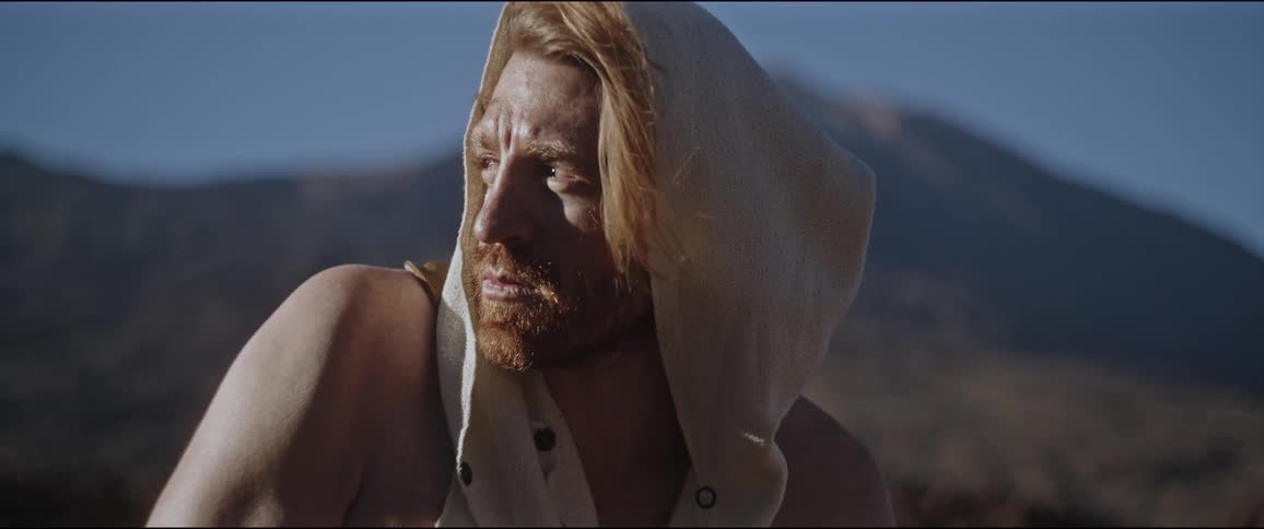 A bearded man with long hair wearing a sleeveless hoodie in front of a mountain