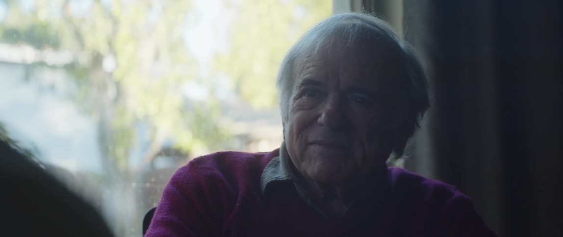 An elderly man sits in a house listening to another person speak
