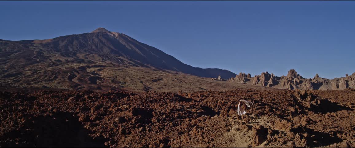 Arid landscape with a large hill in the background under a clear sky and rocky terrain in the foreground with a woman in a dress
