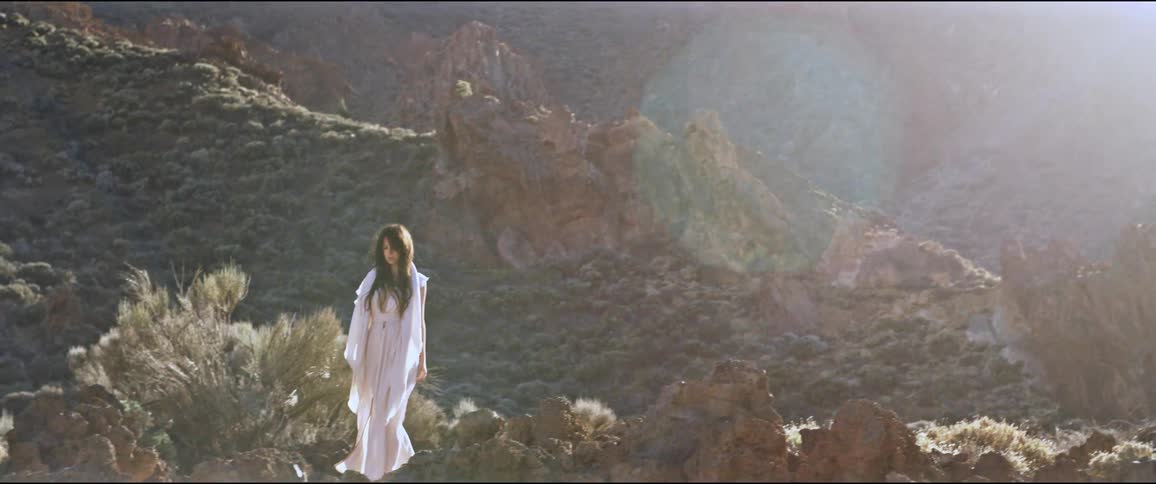 Woman in a white dress walking on rocky terrain with sparse vegetation in sunlight with mountains in the background