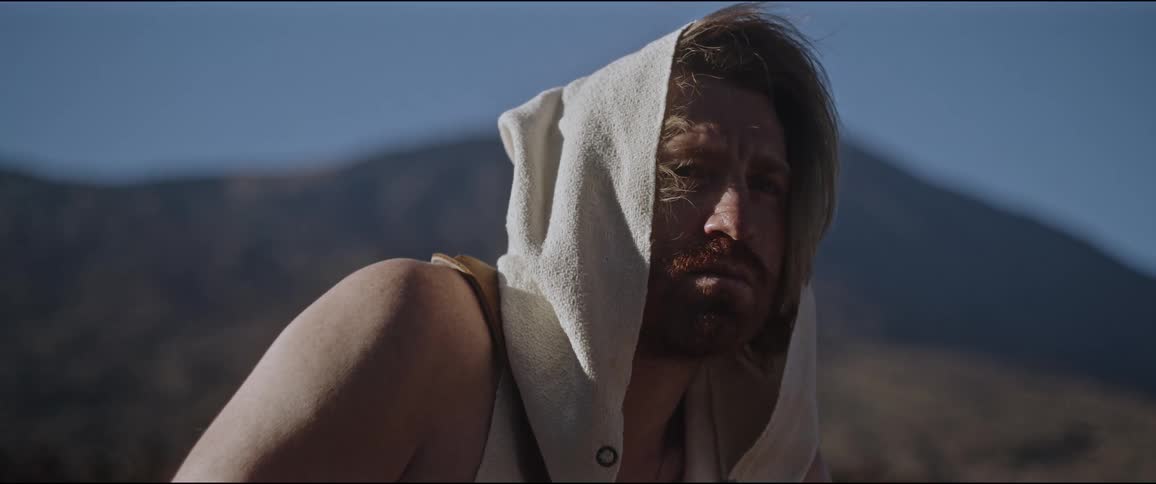 A bearded man with long hair wearing a sleeveless hoodie in front of a mountain