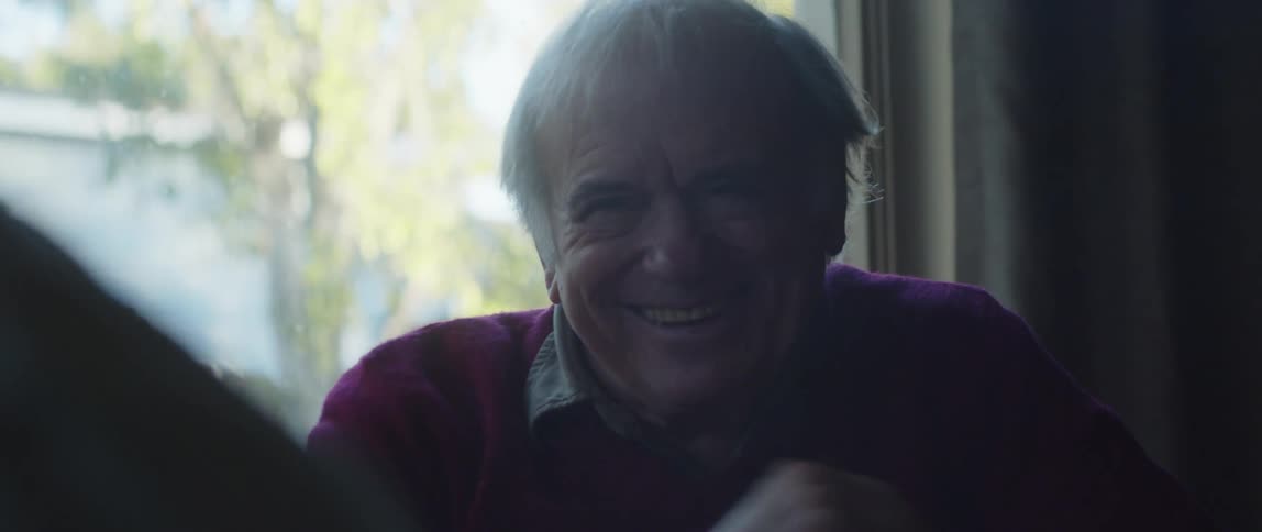 An elderly man smiles and laughs while having a conversation at home