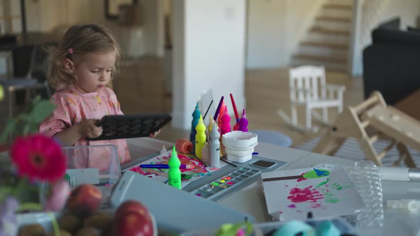 A little girl picks up a piece of plastic near her painting area and looks at it