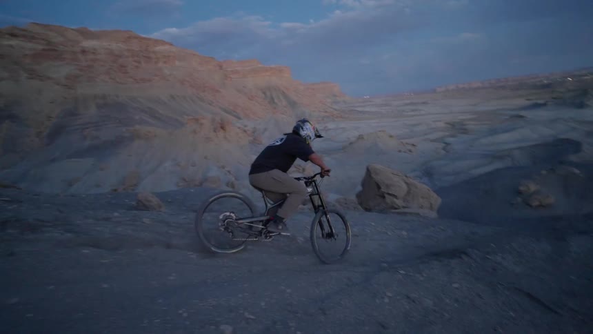Person riding a bicycle on rocky terrain with land formations in the background under a dark blue sky