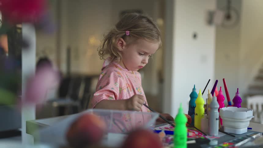 A girl sitting at a table at home uses a brush to paint with water colors
