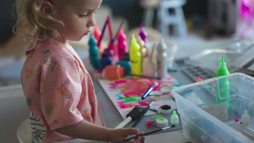 A young girl wearing a pink frock paints on a table