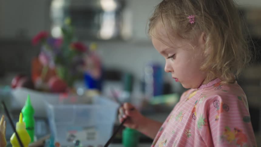 A young girl in a pink top paints a colorful picture on a cluttered table