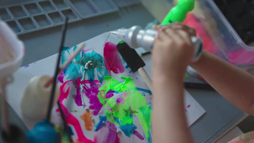 A little girl in a pink top squirts paint out of a bottle and onto her painting