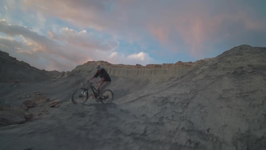 A man bikes down a mountainous terrain riding over a bike jump at dusk