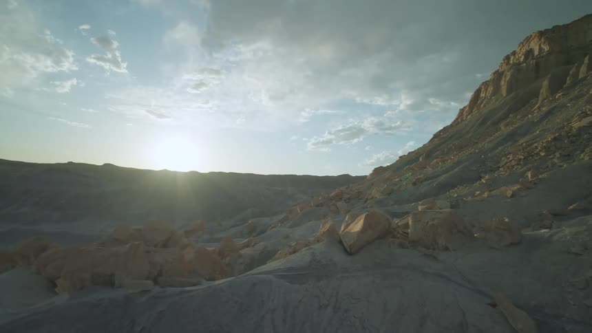 Sun glares from the horizon onto a cliff towering over uneven boulder and rock strewn terrain