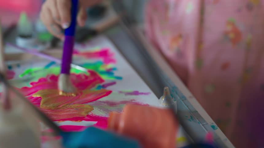Little girl uses a paintbrush to put colored paint on paper on the table