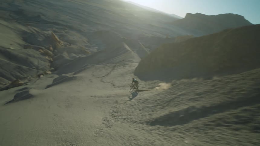 A person riding a bicycle down a narrow dirt trail between tall steep rock formations in the desert