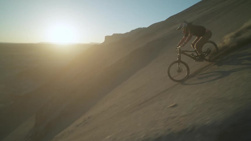 Sun glares from the horizon onto a cyclist riding a bike down a steep dirt mountain slope