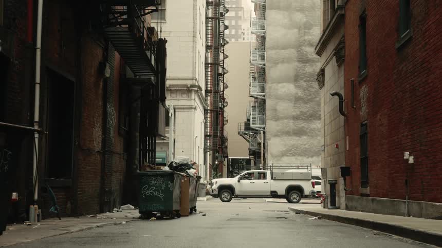 Narrow street between buildings with a dumpster on the left and a white vehicle in the background