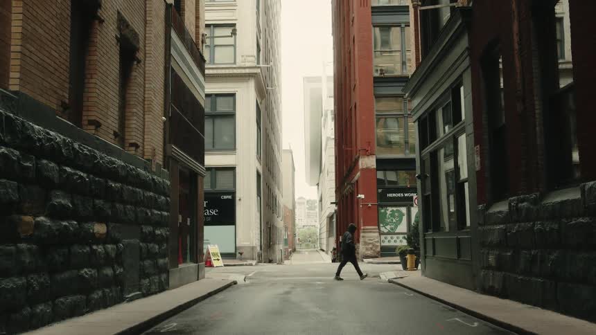 A person crosses an urban street flanked by tall buildings with a clear sky visible at the end of the road