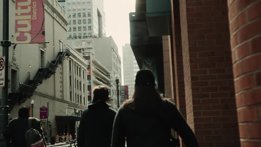 Two people are back to back and walking on a sidewalk and there are buildings right next to them made of bricks
