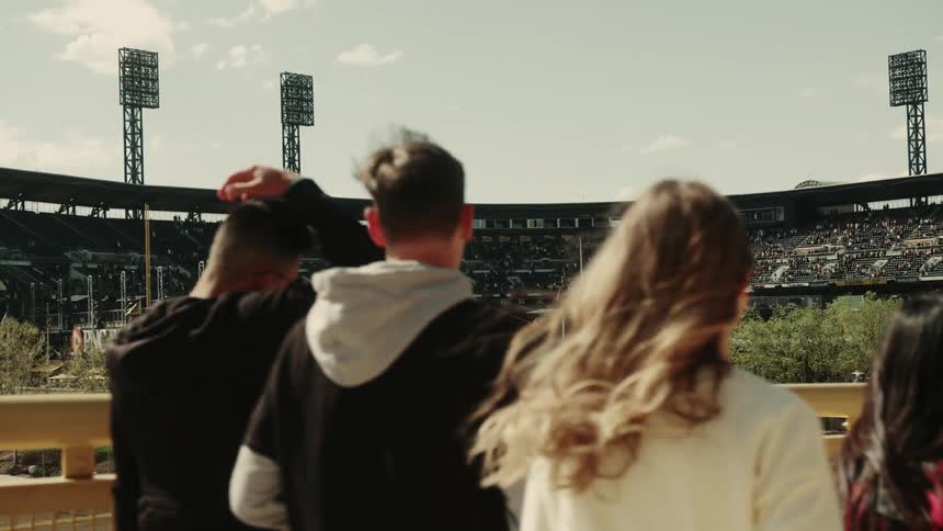 People walk past a rail with a filled sports stadium in the background