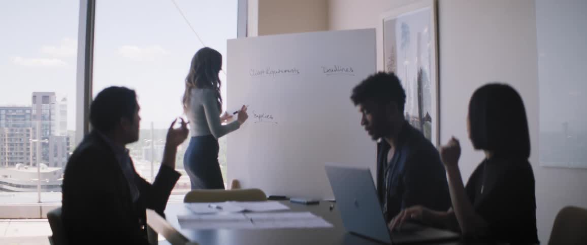 Four people at an office table focused on a woman who is writing on a whiteboard
