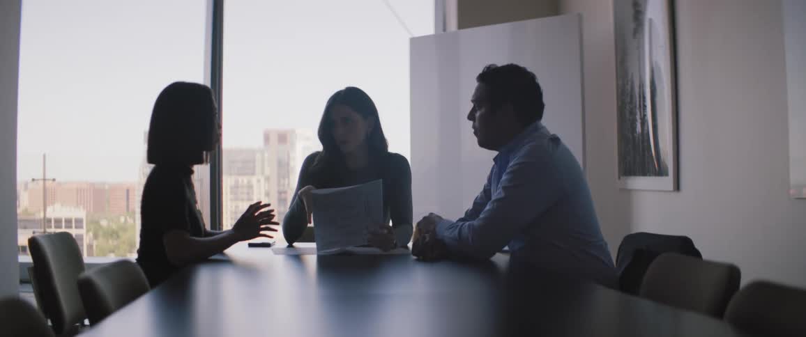 A meeting of three people around a black table and a woman is holding a paper while speaking