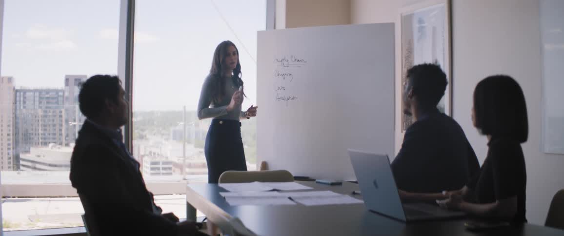 A meeting with three people sitting and one of them is working on a notebook and one person is standing presenting information on a paper