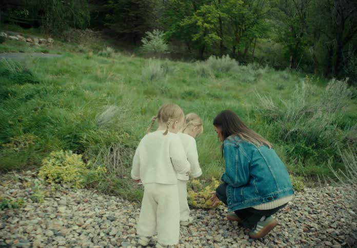 A mother kneels down to pick flowers for her two daughters