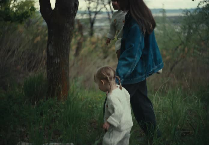 A mother and her two daughters walk through a grassy area near a boulder and tree