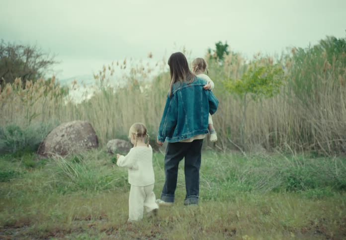 A mother and her daughters walking through a grassy field with tall reeds under an overcast sky
