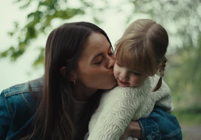 Mother crouches to hug and kiss her daughter while near a hill outdoors