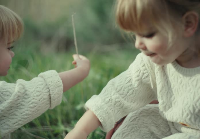 Two girls wearing knitted jumpers playing with blades of green grass
