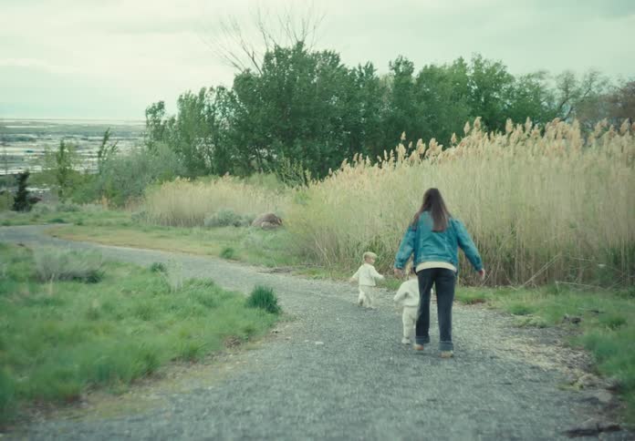A mother and her daughters running on a path near a grassy field with tall reeds under an overcast sky