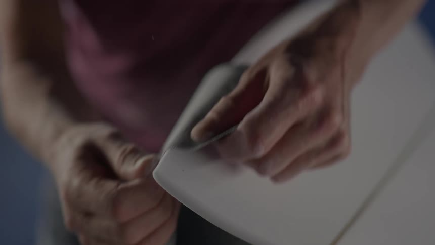 Hands scraping the corner of a surfboard with sandpaper