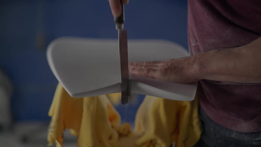 A closeup of a persons hands cutting the tail of a surfboard in a workshop